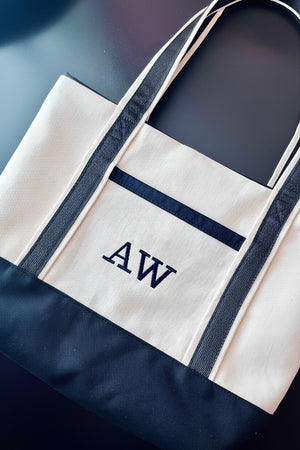 A canvas tote bag with a cream body, dark navy blue trim, and a navy blue bottom panel, featuring the embroidered initials 'AW' on the front pocket.